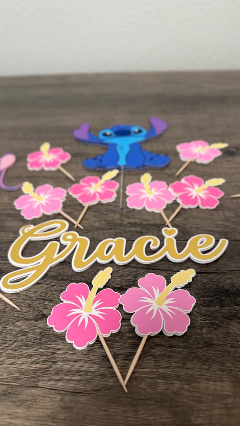 May include: Cake toppers featuring a blue cartoon character and pink hibiscus flowers. The name "Gracie" is written in gold script. The toppers are on wooden sticks, perfect for a themed party.