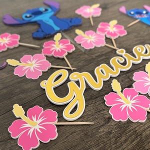 May include: A collection of party decorations featuring cartoon characters and floral accents. Includes blue and purple character cutouts, pink hibiscus flower toppers, and a gold and white name cutout that reads "Gracie".