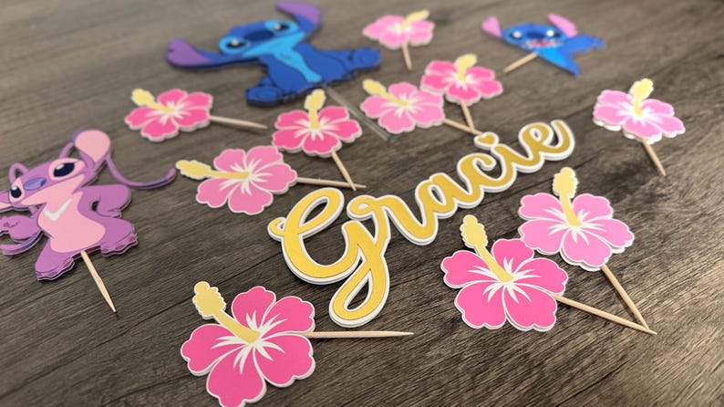 May include: A collection of party decorations featuring cartoon characters and floral accents. Includes blue and purple character cutouts, pink hibiscus flower toppers, and a gold and white name cutout that reads "Gracie".