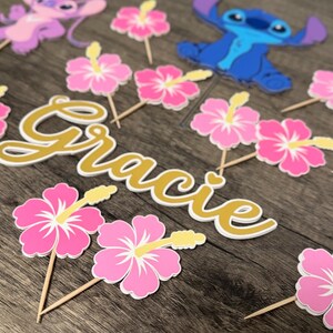May include: A collection of party decorations featuring the name "Gracie" in gold script, along with pink hibiscus flower and cartoon character toppers. The decorations are arranged on a wooden surface.
