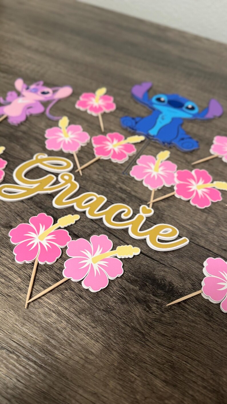 May include: A collection of party decorations featuring the name "Gracie" in gold script, along with pink hibiscus flower and cartoon character toppers. The decorations are arranged on a wooden surface.