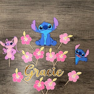 May include: Cake toppers featuring the character Stitch in blue, Angel in pink, and pink hibiscus flowers. The name "Gracie" is written in gold script. Perfect for a themed birthday party.
