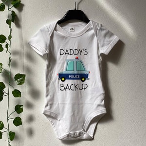 Könnte beinhalten: Weißer Baby-Strampler mit kurzen Ärmeln, auf dem ein blaues und weißes Polizeiauto mit dem Text "Daddy's Backup" abgebildet ist.