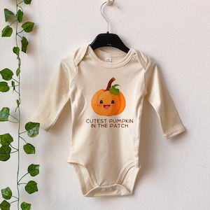 May include: A cream coloured long-sleeved baby onesie with a smiling orange pumpkin graphic and the text "Cutest Pumpkin in the Patch".