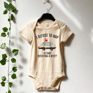 Può includere: Un body da bambino beige chiaro a maniche corte, appeso a una gruccia nera. Il body presenta la scritta "I REFUSE TO NAP" sopra una grafica di un'auto della polizia e la scritta "IS THAT RESISTING A REST?" sotto.