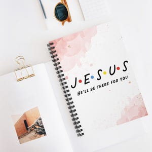 May include: A spiral-bound notebook with a white cover featuring soft pink watercolour splashes and the black text "J E S U S" with colourful dots between letters, followed by the phrase "HE'LL BE THERE FOR YOU" in black capital letters.