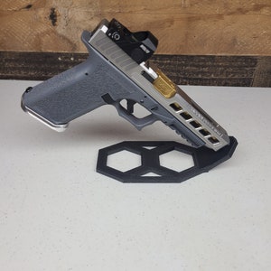 K&ouml;nnte beinhalten: Eine silberne und schwarze Pistole mit einem roten Punktvisier und einer goldenen Akzent auf dem Schlitten. Die Waffe ruht auf einem schwarzen sechseckigen St&auml;nder.