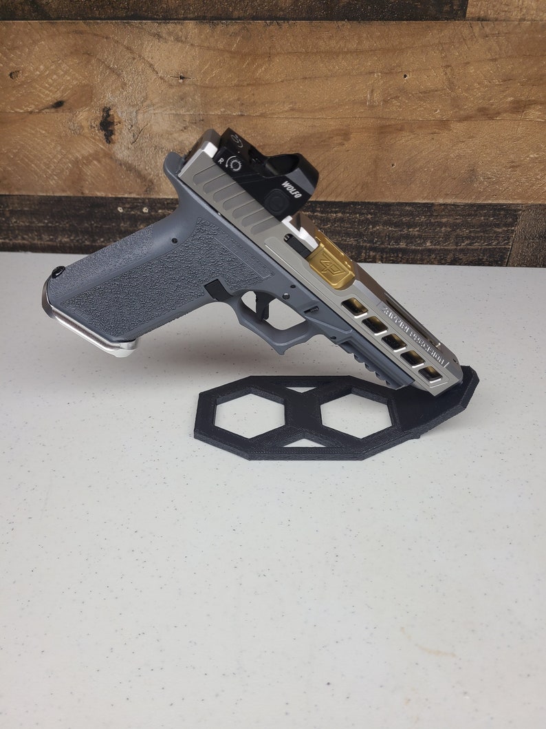K&ouml;nnte beinhalten: Eine silberne und schwarze Pistole mit einem roten Punktvisier und einer goldenen Akzent auf dem Schlitten. Die Waffe ruht auf einem schwarzen sechseckigen St&auml;nder.