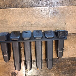 May include: A black plastic magazine holder with six slots for storing magazines. The holder is mounted on a wooden wall.