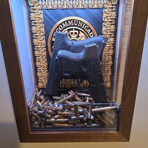 May include: A shadow box display with a wooden frame. Inside, two black handguns are displayed against a backdrop with the word "COMMUNICAD". The bottom of the box is filled with spent bullet casings.