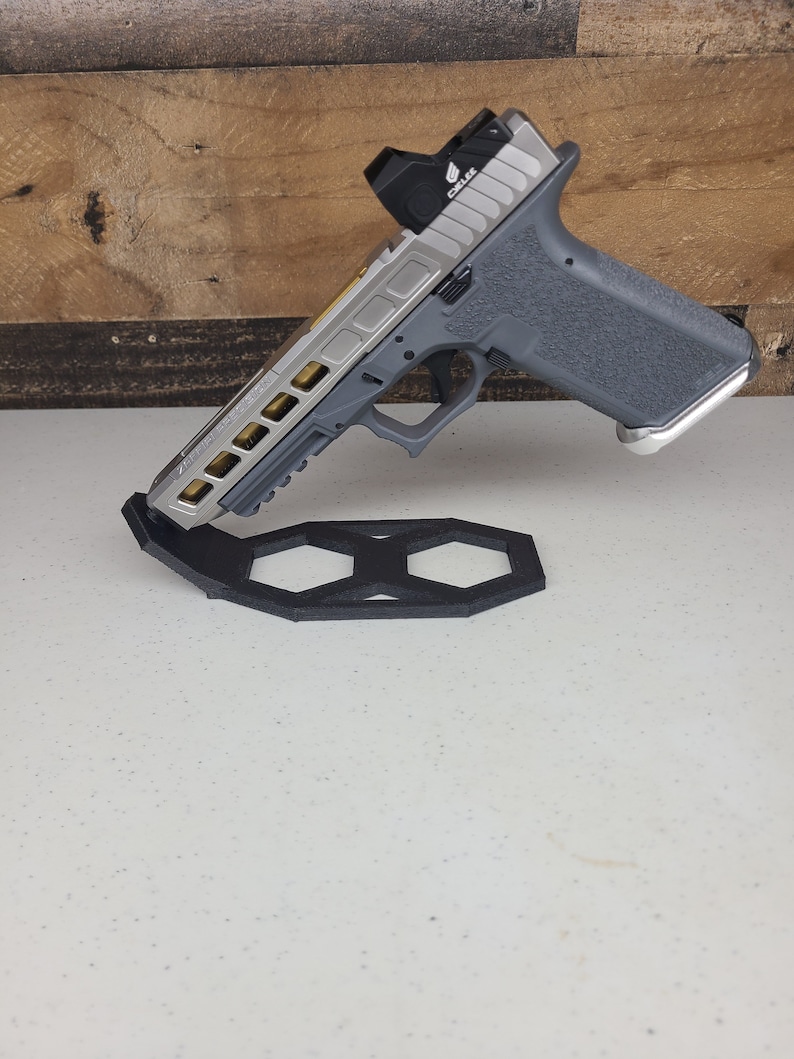K&ouml;nnte beinhalten: Eine silberne und graue Pistole mit einem goldfarbenen Schlitten und einem Rotpunktvisier. Die Waffe steht auf einem schwarzen sechseckigen St&auml;nder.