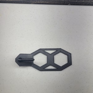 K&ouml;nnte beinhalten: Eine geometrische Form aus schwarzem Kunststoff mit hexagonalem Design. Die Form hat einen kleinen Griff auf der linken Seite.