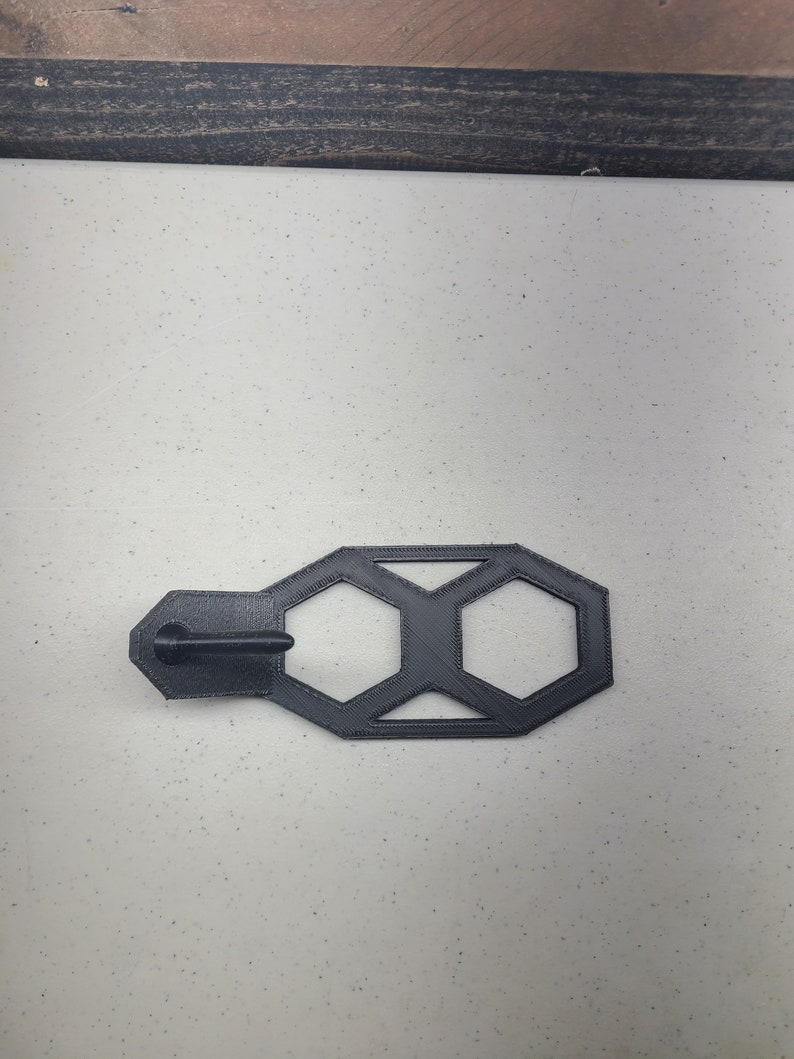 K&ouml;nnte beinhalten: Eine geometrische Form aus schwarzem Kunststoff mit hexagonalem Design. Die Form hat einen kleinen Griff auf der linken Seite.