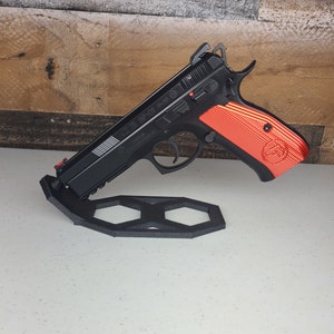 K&ouml;nnte beinhalten: Eine schwarze Pistole mit einem rot-schwarzen Griff wird auf einem schwarzen sechseckigen St&auml;nder pr&auml;sentiert. Die Waffe hat einen silbernen Schlitten mit dem aufgedruckten Text "CZ 75 SP-01".