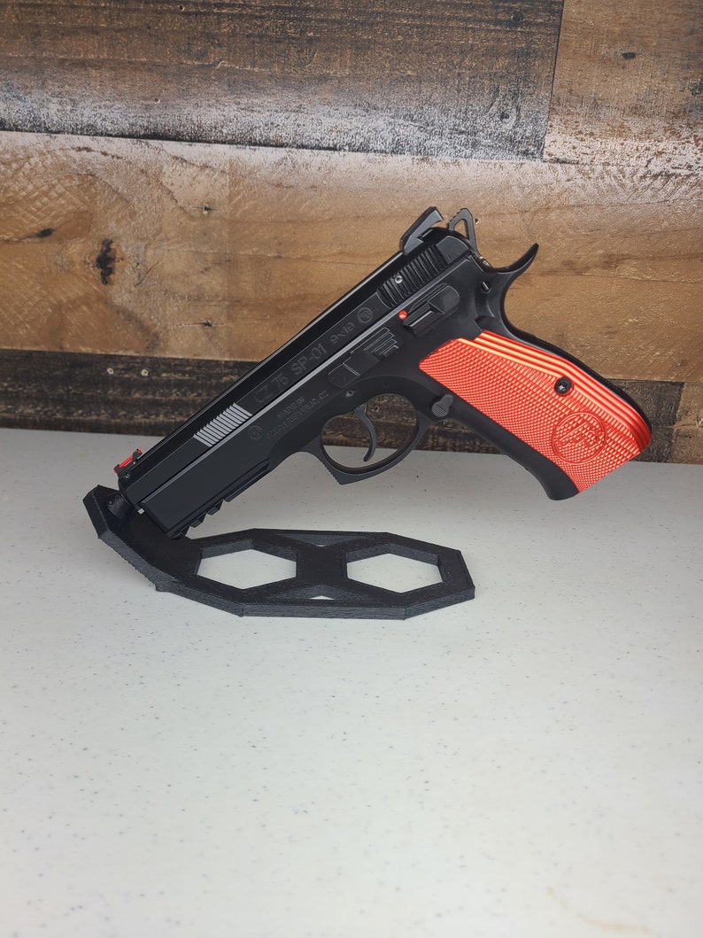 K&ouml;nnte beinhalten: Eine schwarze Pistole mit einem rot-schwarzen Griff wird auf einem schwarzen sechseckigen St&auml;nder pr&auml;sentiert. Die Waffe hat einen silbernen Schlitten mit dem aufgedruckten Text "CZ 75 SP-01".