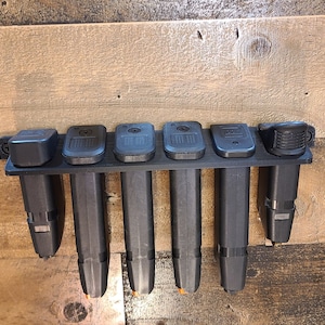 May include: A black plastic magazine holder with six slots for storing magazines. The holder is mounted on a wooden wall.