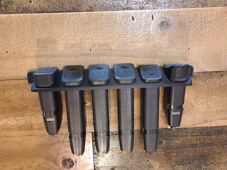 May include: A black plastic magazine holder with six slots for storing magazines. The holder is mounted on a wooden wall.