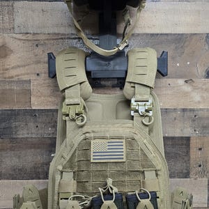 May include: Tan tactical helmet and plate carrier with an American flag patch. The helmet has a mounting system and the carrier has multiple pouches and magazine holders. The gear is displayed against a wooden background.