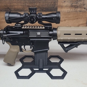 May include: A black and tan rifle with a scope mounted on top. The rifle is resting on a black hexagonal stand. The scope has a black body and a silver dial. The rifle has a black handguard and a tan stock.
