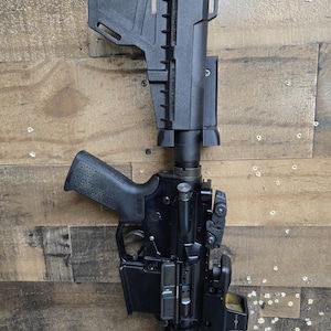May include: Black rifle with a black stock, grip, and various attachments. The rifle is set against a wooden background. The rifle has a red dot sight and a magazine.