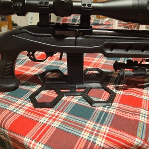 May include: A black rifle with a scope mounted on top, resting on a black hexagonal stand. The rifle is on a red and green plaid tablecloth.