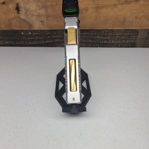 K&ouml;nnte beinhalten: Eine silberne und goldene Pistole mit einem schwarzen Rotpunktvisier, das oben angebracht ist. Die Waffe ist auf einem schwarzen St&auml;nder montiert.