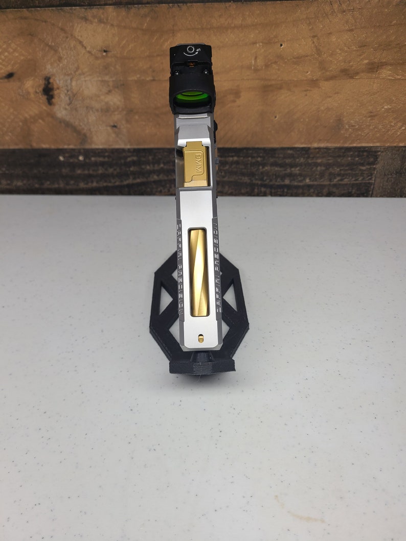 K&ouml;nnte beinhalten: Eine silberne und goldene Pistole mit einem schwarzen Rotpunktvisier, das oben angebracht ist. Die Waffe ist auf einem schwarzen St&auml;nder montiert.