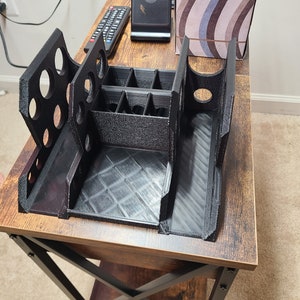 May include: A black plastic organizer with multiple compartments and slots. The organizer is sitting on a wooden table with a black metal frame.