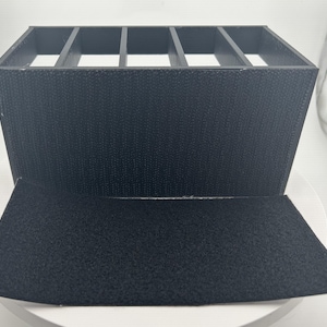 May include: A black organizer with multiple compartments, constructed from a textured material. The organizer has a rectangular shape with an open top and a flap that folds down. The compartments are divided by vertical dividers.