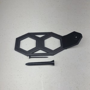 K&ouml;nnte beinhalten: Ein schwarzer 3D-gedruckter Wandhalter mit zwei sechseckigen &Ouml;ffnungen und einem schwarzen Lederriemen. Der Halter ist so konzipiert, dass er zwei Gegenst&auml;nde, wie z. B. Schl&uuml;ssel oder Schmuck, aufnehmen kann. Eine schwarze Schraube ist im Lieferumfang enthalten.