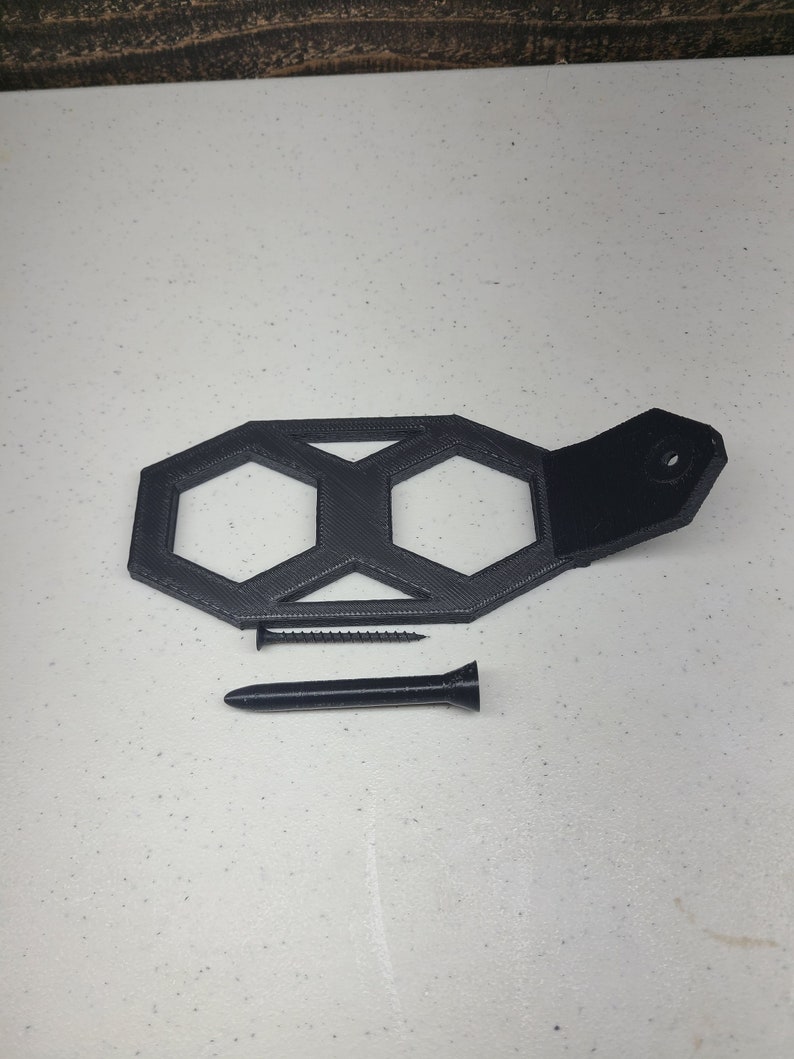K&ouml;nnte beinhalten: Ein schwarzer 3D-gedruckter Wandhalter mit zwei sechseckigen &Ouml;ffnungen und einem schwarzen Lederriemen. Der Halter ist so konzipiert, dass er zwei Gegenst&auml;nde, wie z. B. Schl&uuml;ssel oder Schmuck, aufnehmen kann. Eine schwarze Schraube ist im Lieferumfang enthalten.