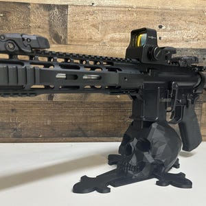 May include: Black rifle with a red dot sight, mounted on a black skull-shaped stand. The rifle has a tactical rail system and a suppressor. The background is a wooden wall.