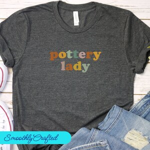 May include: A dark gray t-shirt with the text "pottery lady" in colorful letters. The letters are in different shades of orange, brown, green, and pink.