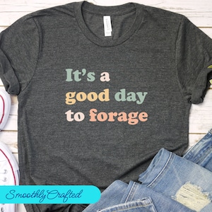May include: A dark gray t-shirt with the text "It's a good day to forage" printed in colorful letters. The text is in a retro style font.