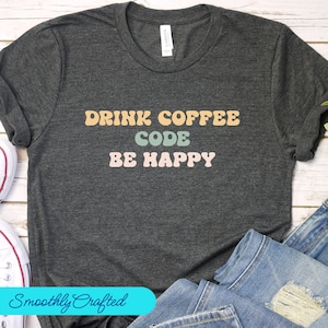 Puede incluir: Una camiseta gris oscuro con el texto "Drink Coffee Code Be Happy" en una fuente retro con letras de diferentes colores. El texto está centrado en la camiseta.