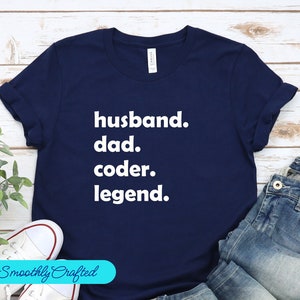 May include: A navy blue t-shirt with the words "husband. dad. coder. legend." printed in white. The sleeves are rolled up. The image also shows blue jeans. A light blue banner with "Smoothly Crafted" is in the bottom left corner.