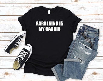 Può includere: Maglietta nera con testo bianco che recita "Gardening is my cardio". La maglietta è appoggiata su una superficie di legno bianco con un paio di scarpe da ginnastica nere e jeans blu.