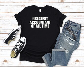 Könnte beinhalten: Schwarzes T-Shirt mit weißem Text, der "Greatest Accountant of All Time" lautet. Das Shirt liegt flach auf einer Holzoberfläche mit einem Paar schwarzer Turnschuhe und einer blauen Jeans.