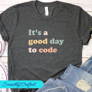 Puede incluir: Una camiseta gris oscuro con el texto "It's a good day to code" en letras de colores. El texto está en un estilo de fuente retro.