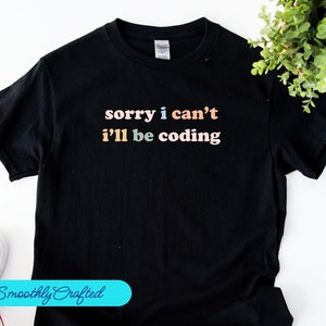 Op de afbeelding: Zwart T-shirt met de tekst "sorry i can't i'll be coding" in regenboogkleuren.