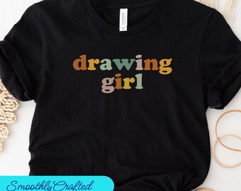 Può includere: T-shirt nero con la scritta "drawing girl" in un font retrò. Il testo è nei toni del marrone, giallo, verde e rosa. La maglietta è realizzata in un materiale morbido.