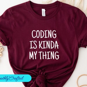 Könnte beinhalten: Burgunderfarbenes T-Shirt mit weißem Text, der "Coding is kinda my thing" lautet.
