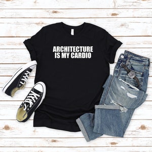 May include: Black t-shirt with white text that reads "Architecture is my cardio". The shirt is laid flat on a white wood surface with a pair of black sneakers and blue jeans.