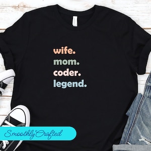May include: Black t-shirt with the text "wife. mom. coder. legend." in pastel colours. The text is stacked vertically in the centre of the shirt. The shirt is on a wooden surface with a pair of black trainers and a pair of blue jeans.
