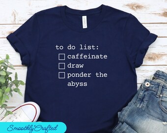 Può includere: T-shirt blu navy con la scritta bianca "to do list: caffeinate, draw, ponder the abyss". La maglietta ha le maniche arrotolate ed è abbinata a jeans blu e scarpe da ginnastica bianche.
