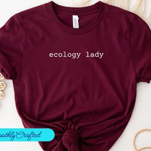Può includere: Una maglietta color bordeaux con la scritta "ecology lady" stampata in bianco sul davanti.