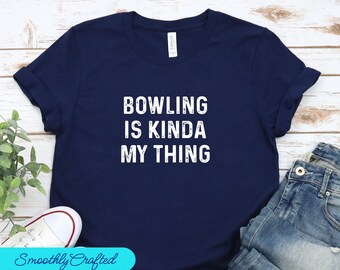 May include: A navy blue t-shirt with white text that reads "Bowling is kinda my thing". The shirt is folded and displayed on a wooden surface with a pair of white sneakers and blue jeans.
