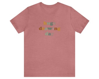 Può includere: Una t-shirt color rosa tenue con il testo "huge drawing day" in lettere colorate.