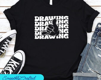 Può includere: T-shirt nera con la parola "DRAWING" impilata quattro volte in bianco, con una grafica di una mano dei cartoni animati. La maglietta è abbinata a jeans blu e scarpe da ginnastica nere.