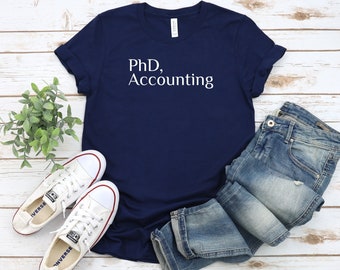 Könnte beinhalten: Ein marineblaues T-Shirt mit dem Text "PhD, Accounting" in weißen Buchstaben. Das Shirt liegt flach auf einer weißen Holzoberfläche mit einem Paar weißen Sneakers und einer blauen Jeans.